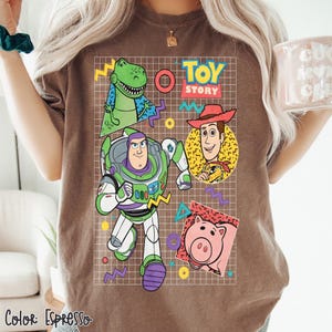 May include: Espresso-colored t-shirt featuring a graphic of Toy Story characters: Rex, Buzz Lightyear, Woody, and Hamm. The design includes the words "Toy Story" and colorful geometric shapes. The shirt's color is labeled "Espresso" at the bottom.
