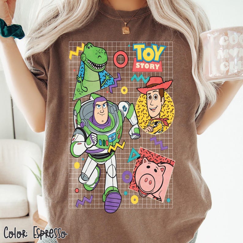 May include: Espresso-colored t-shirt featuring a graphic of Toy Story characters: Rex, Buzz Lightyear, Woody, and Hamm. The design includes the words "Toy Story" and colorful geometric shapes. The shirt's color is labeled "Espresso" at the bottom.