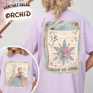 May include: Orchid Comfort Color t-shirt featuring a graphic of a musical score with the text "Let It Go" and illustrations of characters from a popular animated film. The design includes a snowflake and floral accents.
