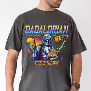 May include: A dark gray t-shirt with a retro-style graphic of a Mandalorian character holding a small green creature. The graphic includes the text "The Dadalorian" and "This is the Way".
