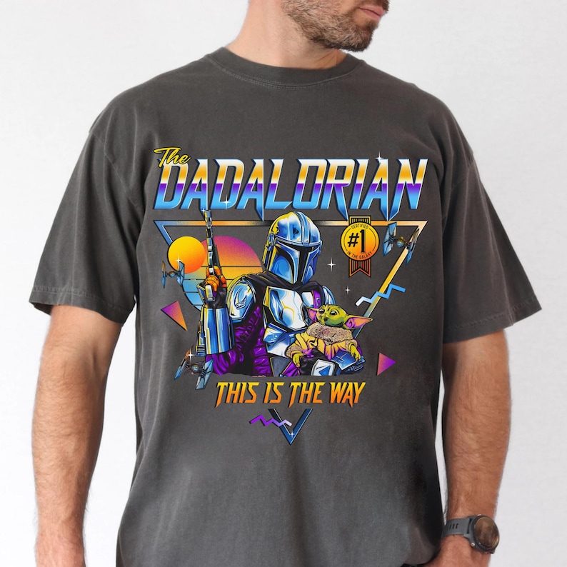 May include: A dark gray t-shirt with a retro-style graphic of a Mandalorian character holding a small green creature. The graphic includes the text "The Dadalorian" and "This is the Way".