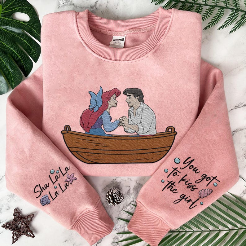 May include: Pink sweatshirt featuring a graphic of Ariel and Prince Eric in a boat. The sleeves have text that reads "Sha La La La La" and "You got to kiss the girl".
