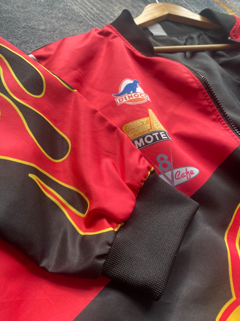 May include: Red and black bomber jacket with a large flame design on the front. The jacket has a zipper closure and features several patches, including a Dinoco logo, a Cozy Cone Motel sign, and a cafe sign.