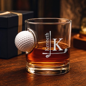 May include: A clear glass tumbler featuring an embedded white golf ball on the side. The glass is etched with the name Kenneth, a large letter K, and two crossed golf clubs. The glass is filled with an amber-colored liquid.