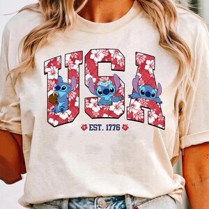 May include: A cream-colored t-shirt featuring the word "USA" in red letters filled with a floral pattern. The letters contain cartoon characters. Below the letters is the text "EST. 1776" with floral accents.