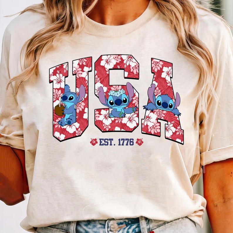 May include: A cream-colored t-shirt featuring the word "USA" in red letters filled with a floral pattern. The letters contain cartoon characters. Below the letters is the text "EST. 1776" with floral accents.