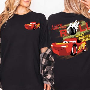 Puede incluir: Camiseta negra con un dise&ntilde;o de coche de dibujos animados rojo. La parte delantera tiene un peque&ntilde;o gr&aacute;fico de coche, mientras que la parte trasera muestra un coche m&aacute;s grande con el texto "I LIKE HOW YOU ROLL" y "WALT DISNEY WORLD".