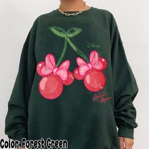 Op de afbeelding: Boszwarte sweatshirt met een Minnie Mouse kersendesign. De afbeelding bevat rode kersen met roze strikken, groene bladeren en het Disney-logo. De woorden "Minnie Mouse" zijn cursief geschreven.