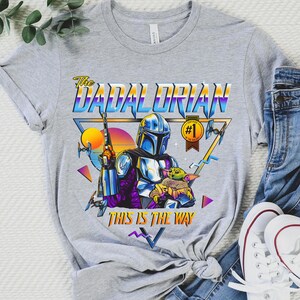May include: A gray t-shirt with a retro-style graphic of a Mandalorian character holding a child. The graphic includes the text "The Dadalorian" and "This is the way".