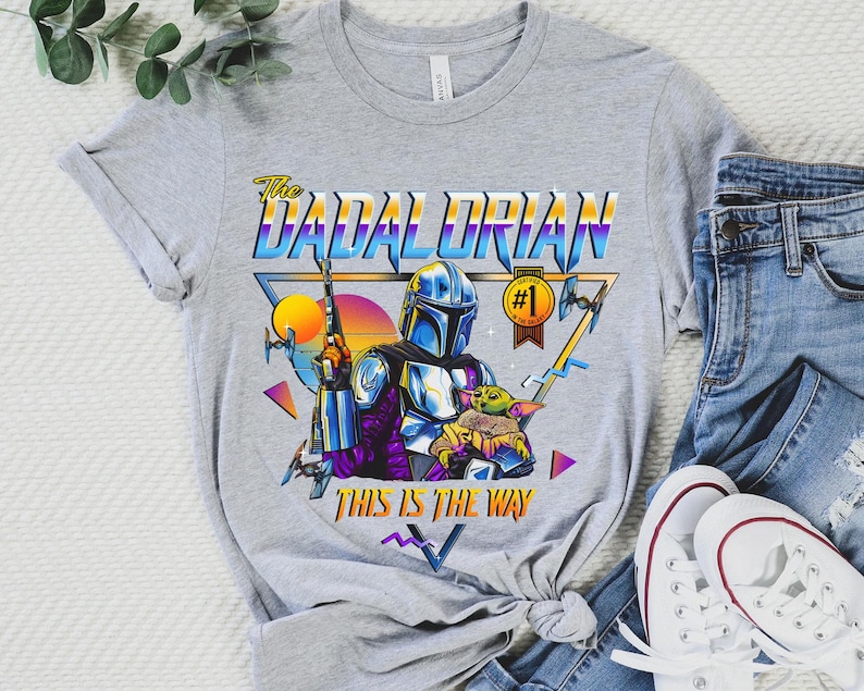 May include: A gray t-shirt with a retro-style graphic of a Mandalorian character holding a child. The graphic includes the text "The Dadalorian" and "This is the way".