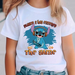 Puede incluir: Camiseta blanca con un personaje azul Stitch sosteniendo un mando de videojuego. El texto "Player 2 has entered the game" est&aacute; impreso en la camiseta.
