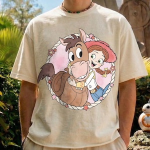 May include: Beige t-shirt featuring a circular graphic of a cartoon horse hugging a cowgirl. The horse is brown, and the cowgirl has red hair and a pink hat. The graphic is surrounded by a white rope border with pink stars.