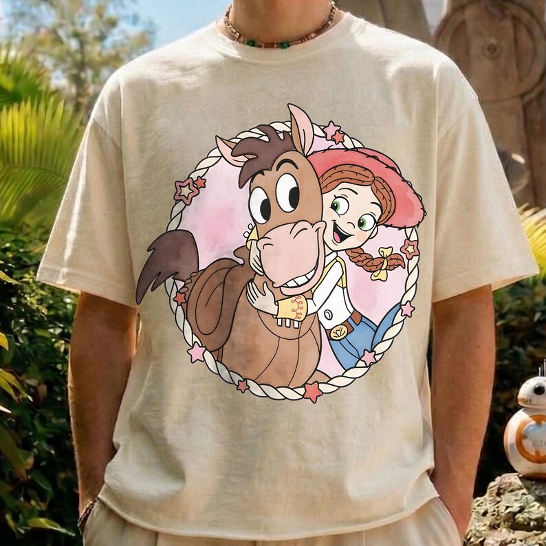 May include: Beige t-shirt featuring a circular graphic of a cartoon horse hugging a cowgirl. The horse is brown, and the cowgirl has red hair and a pink hat. The graphic is surrounded by a white rope border with pink stars.
