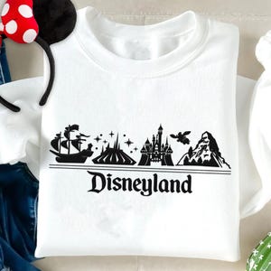 Op de afbeelding: Witte crewneck sweatshirt met een zwarte Disneyland-afbeelding. De afbeelding toont een schip, Space Mountain, een kasteel, een vliegend wezen en een bergketen, met het woord "Disneyland" eronder.