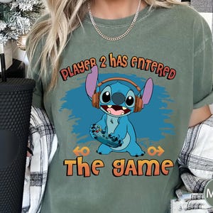 Puede incluir: Una camiseta verde con un personaje azul Stitch sosteniendo un mando de videojuego. El texto "Player 2 has entered the game" est&aacute; impreso en la camiseta.