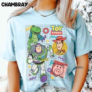 May include: Light blue t-shirt featuring a colorful graphic of characters from the animated film "Toy Story." The design includes Buzz Lightyear, Woody, Rex, Hamm, and the words "Toy Story" in a retro style. The shirt has short sleeves.