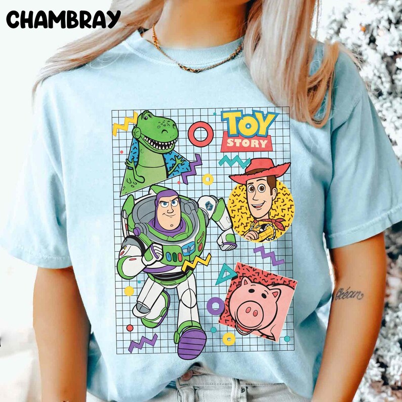 May include: Light blue t-shirt featuring a colorful graphic of characters from the animated film "Toy Story." The design includes Buzz Lightyear, Woody, Rex, Hamm, and the words "Toy Story" in a retro style. The shirt has short sleeves.