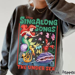 Pu&ograve; includere: Felpa grigio scuro con una grafica colorata di "Singalong Songs" della Disney. Il design include Ariel la sirena, pesci e altri personaggi de "La Sirenetta". Il testo "THE UNDER SEA" &egrave; in basso.