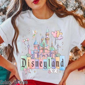 Puede incluir: Camiseta blanca con un gr&aacute;fico colorido de Disneyland. El dise&ntilde;o incluye un castillo, Campanilla, globos y la palabra "Disneyland" en una fuente pastel. Tambi&eacute;n se incluye un lazo y un gesto de la mano en forma de coraz&oacute;n.