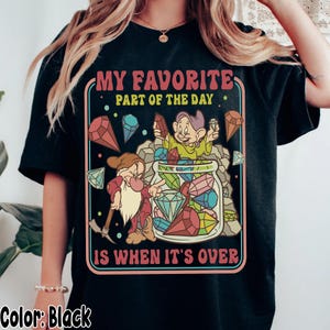 Puede incluir: Camiseta negra con un gráfico colorido que presenta dos enanos de dibujos animados y un tarro lleno de gemas. El texto "MY FAVORITE PART OF THE DAY IS WHEN IT'S OVER" se muestra en una fuente retro. La camiseta es de corte holgado.