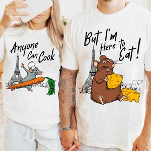 Puede incluir: Dos camisetas blancas con ilustraciones de ratas de dibujos animados y la Torre Eiffel. La camiseta izquierda muestra una rata gris con gorro de chef sosteniendo una zanahoria con el texto Anyone Can Cook. La derecha muestra una rata marrón con queso y el texto But I'm Here to Eat!