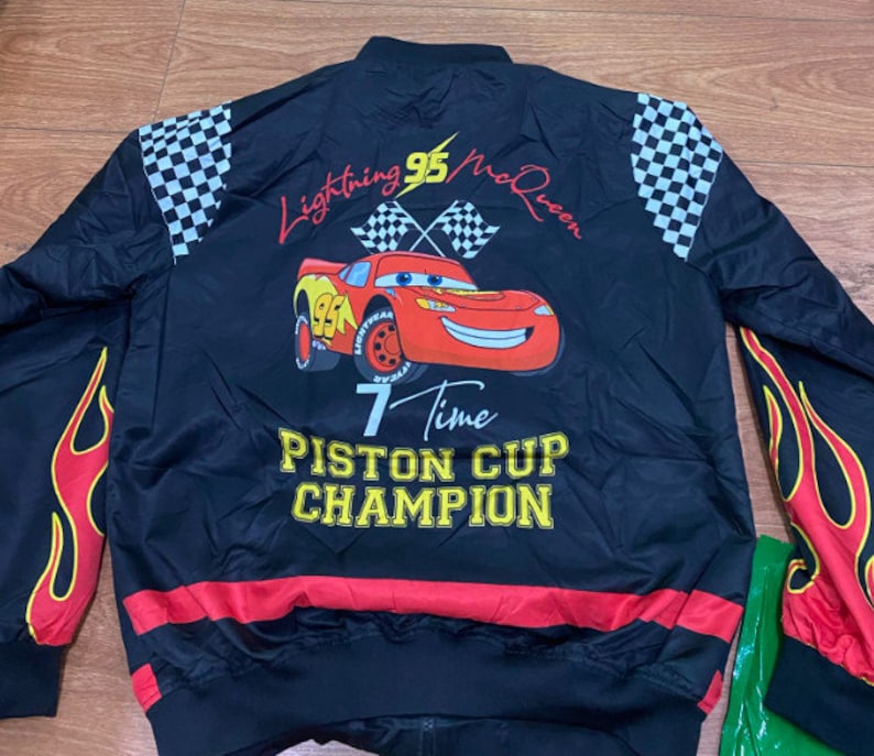 May include: Black bomber jacket with a red, yellow, and black checkered pattern on the sleeves. The jacket features a large graphic of Lightning McQueen from the Cars movie, with the text "Lightning 95 McQueen", "7 Time Piston Cup Champion", and "Piston Cup Champion".