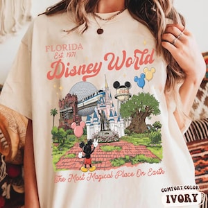 Puede incluir: Camiseta de color marfil con un gr&aacute;fico colorido de los lugares emblem&aacute;ticos de Disney World, incluyendo el castillo de Cenicienta, Spaceship Earth y Mickey Mouse. El texto incluye "FLORIDA Est. 1971" y "Disney World". La frase "The Most Magical Place On Earth" tambi&eacute;n est&aacute; impresa.