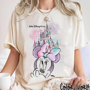 Pode incluir: Camiseta cor marfim com um desenho da Minnie Mouse, um castelo e fogos de artif&iacute;cio. O texto "Walt Disney World" est&aacute; acima do castelo. O desenho inclui um grande la&ccedil;o e &eacute; delineado em preto. As palavras "Color: Ivory" est&atilde;o na parte inferior.