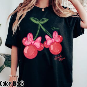 Op de afbeelding: Zwart T-shirt met een Minnie Mouse kers ontwerp. De afbeelding toont twee rode kersen met roze strikken, groene bladeren en het Disney-logo. De woorden "Minnie Mouse" zijn ook aanwezig.