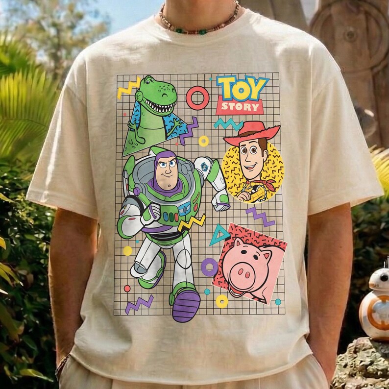May include: Cream-colored t-shirt featuring a graphic of characters from the animated film "Toy Story." The design includes Buzz Lightyear, Woody, Rex, and Hamm, set against a grid background with colorful geometric shapes. The words "TOY STORY" are also visible.