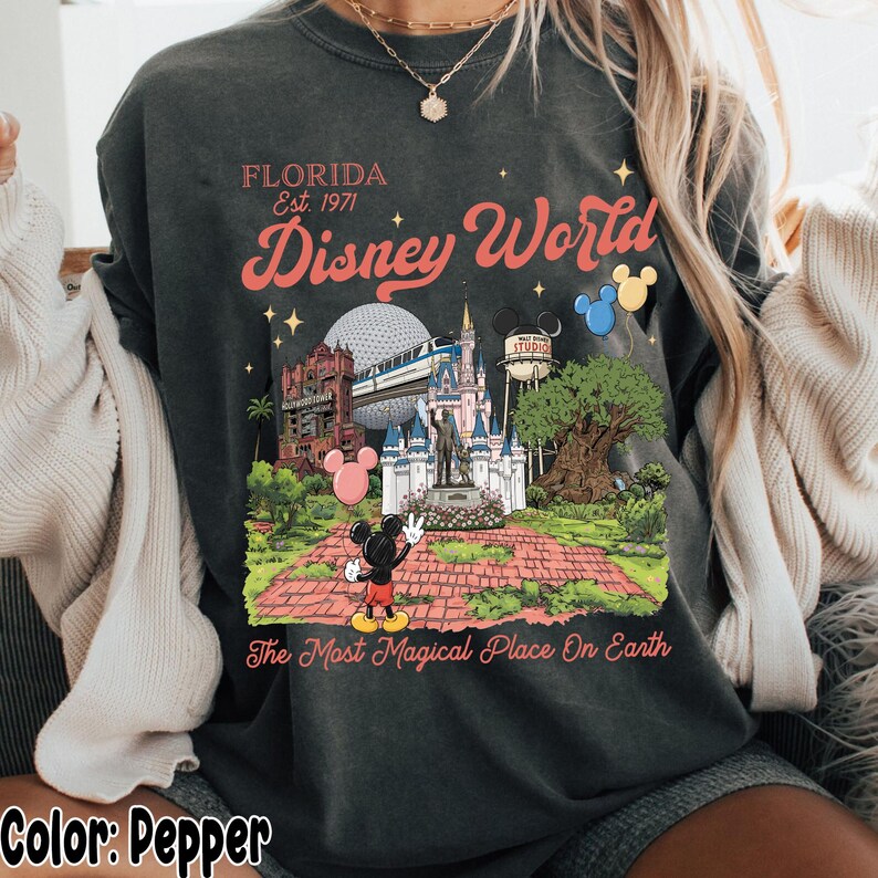 Puede incluir: Sudadera color pimienta con un colorido gr&aacute;fico de Disney World. El dise&ntilde;o incluye el texto "FLORIDA Est. 1971" y "Disney World" con ilustraciones de lugares emblem&aacute;ticos del parque y Mickey Mouse. Tambi&eacute;n incluye la frase "The Most Magical Place On Earth".