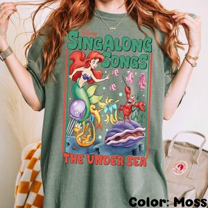 Pu&ograve; includere: T-shirt verde muschio con una grafica colorata de "La Sirenetta" di Disney. Il design include Ariel, Flounder, Sebastian e altri personaggi con il testo "Singalong Songs The Under Sea."