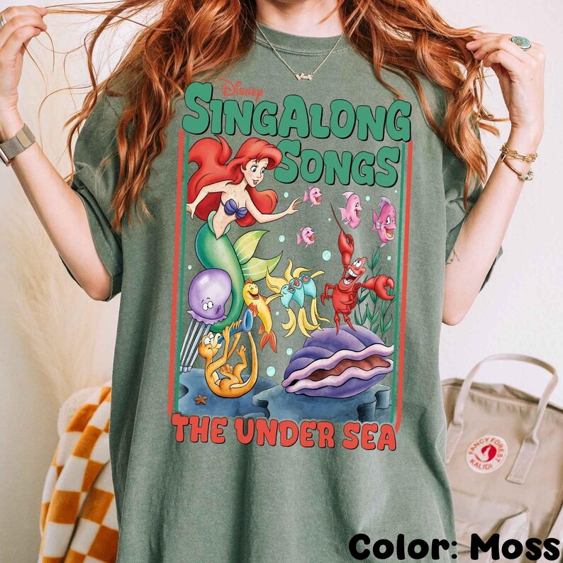 Pu&ograve; includere: T-shirt verde muschio con una grafica colorata de "La Sirenetta" di Disney. Il design include Ariel, Flounder, Sebastian e altri personaggi con il testo "Singalong Songs The Under Sea."