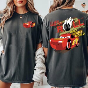 Puede incluir: Dos camisetas de color gris carb&oacute;n con un gr&aacute;fico de un coche de carreras rojo de dibujos animados. Una camiseta tiene el coche en la parte delantera, la otra tiene el coche y el texto "I LIKE HOW YOU ROLL" y "WALT DISNEY WORLD".
