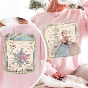 May include: Pink sweatshirt featuring a colorful illustration of Elsa and Anna from Frozen, set against a musical score background. The design includes a snowflake and the text "Let It Go".
