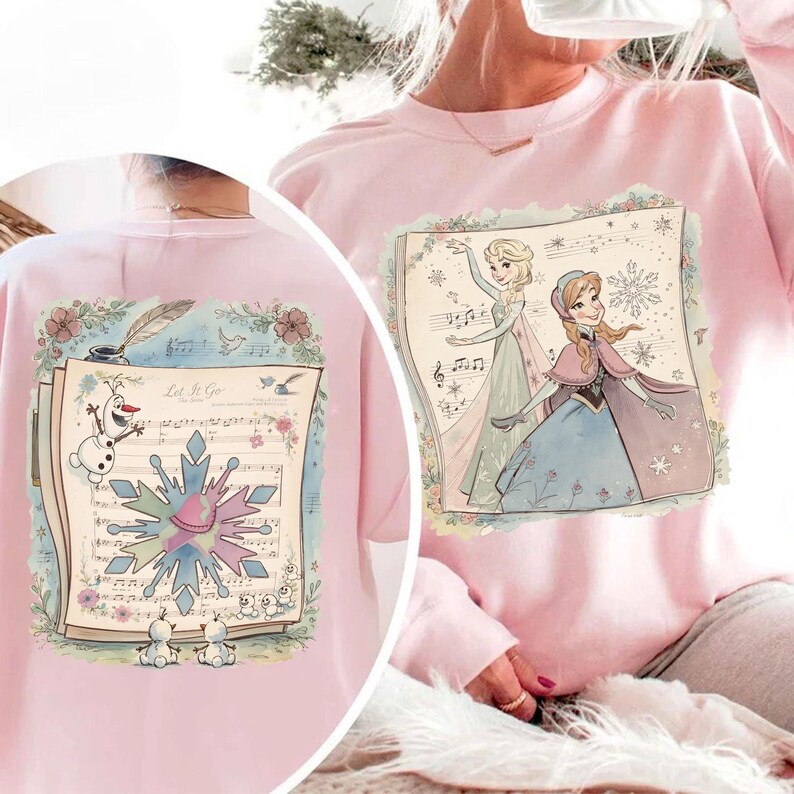 May include: Pink sweatshirt featuring a colorful illustration of Elsa and Anna from Frozen, set against a musical score background. The design includes a snowflake and the text "Let It Go".