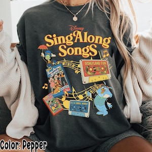 May include: Pepper-colored sweatshirt featuring the text "Disney Sing Along Songs" in yellow. The design includes cartoon characters, cassette tapes, and musical notes. The sweatshirt has a vintage, distressed look.