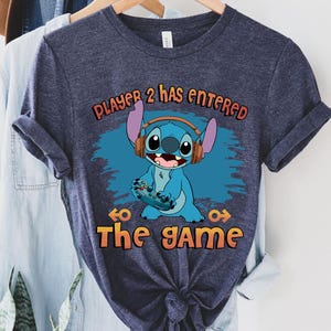 Puede incluir: Una camiseta azul marino con un personaje azul Stitch que lleva auriculares y sostiene un mando de videojuego. El texto "Player 2 has entered the game" est&aacute; impreso en la camiseta.