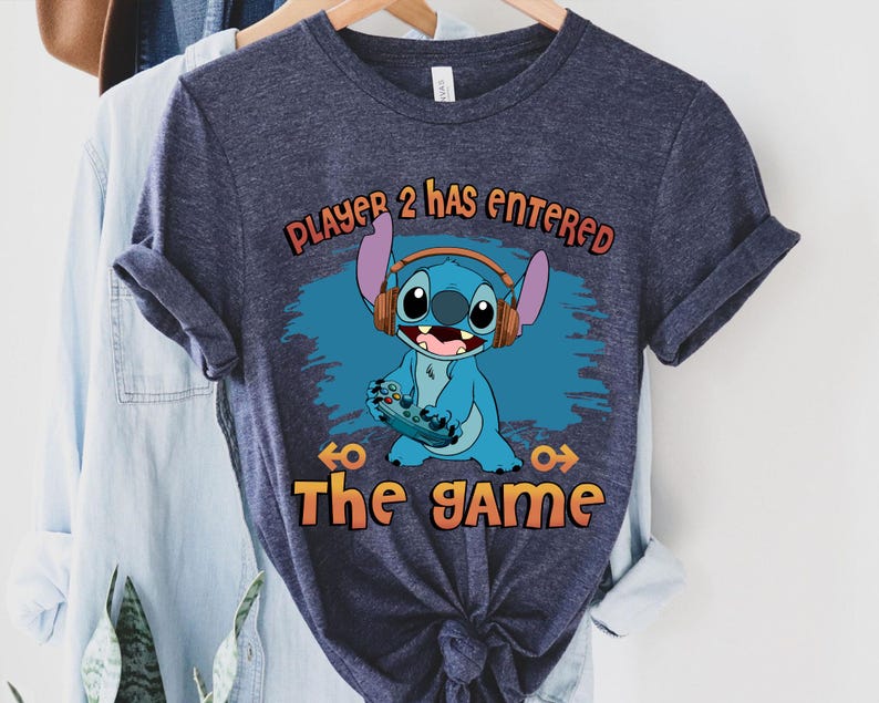 Puede incluir: Una camiseta azul marino con un personaje azul Stitch que lleva auriculares y sostiene un mando de videojuego. El texto "Player 2 has entered the game" est&aacute; impreso en la camiseta.