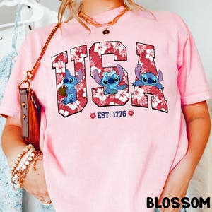 May include: Pink t-shirt with the word "USA" in a floral design, featuring cartoon characters. The text "EST. 1776" is below the word "USA". The word "BLOSSOM" is at the bottom right of the shirt.