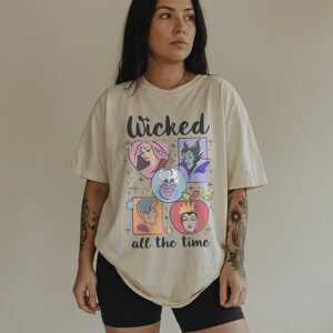 K&ouml;nnte beinhalten: Cremefarbenes T-Shirt mit dem Wort "Wicked" in schwarzer Schrift &uuml;ber einer Grafik von Cartoon-B&ouml;sewichten. Die Grafik enth&auml;lt Bilder eines Herzens, einer lila-h&auml;utigen Frau, einer Kristallkugel und eines Apfels. Der Text "all the time" steht darunter.