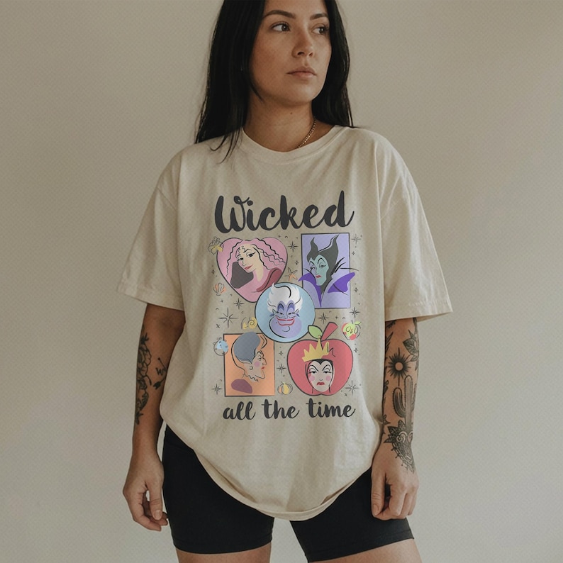 K&ouml;nnte beinhalten: Cremefarbenes T-Shirt mit dem Wort "Wicked" in schwarzer Schrift &uuml;ber einer Grafik von Cartoon-B&ouml;sewichten. Die Grafik enth&auml;lt Bilder eines Herzens, einer lila-h&auml;utigen Frau, einer Kristallkugel und eines Apfels. Der Text "all the time" steht darunter.