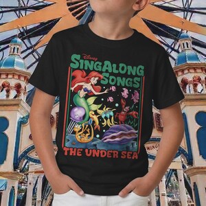Pu&ograve; includere: T-shirt nera con una grafica colorata de "La Sirenetta" della Disney. Il design include Ariel, varie creature marine e il testo "Sing Along Songs: The Under Sea."