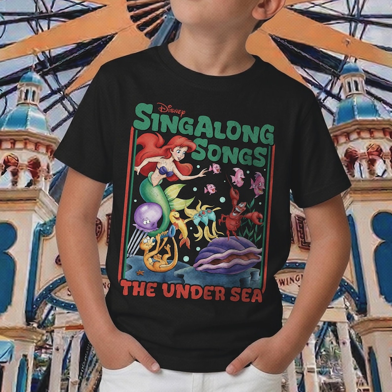 Pu&ograve; includere: T-shirt nera con una grafica colorata de "La Sirenetta" della Disney. Il design include Ariel, varie creature marine e il testo "Sing Along Songs: The Under Sea."
