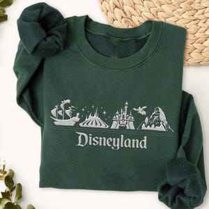Op de afbeelding: Een donkergroene sweatshirt met een wit geborduurd Disneyland-ontwerp. Het ontwerp omvat een schip, berg, kasteel en het woord "Disneyland". De sweatshirt heeft een ronde hals en lange mouwen.