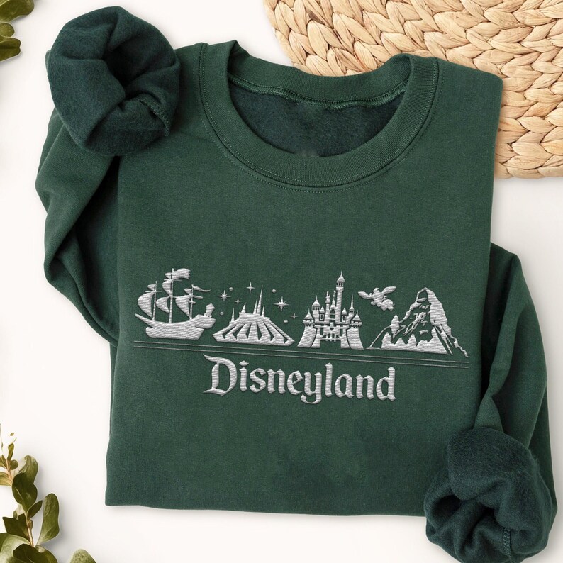 Op de afbeelding: Een donkergroene sweatshirt met een wit geborduurd Disneyland-ontwerp. Het ontwerp omvat een schip, berg, kasteel en het woord "Disneyland". De sweatshirt heeft een ronde hals en lange mouwen.