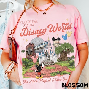 Puede incluir: Camiseta rosa claro con un gr&aacute;fico de Disney World, que incluye el castillo de Cenicienta, el Hollywood Tower Hotel y Mickey Mouse. El texto "Florida Est. 1971 Disney World" est&aacute; impreso encima del gr&aacute;fico. La frase "The Most Magical Place On" est&aacute; impresa debajo.