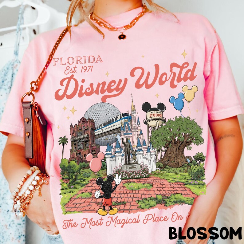 Puede incluir: Camiseta rosa claro con un gr&aacute;fico de Disney World, que incluye el castillo de Cenicienta, el Hollywood Tower Hotel y Mickey Mouse. El texto "Florida Est. 1971 Disney World" est&aacute; impreso encima del gr&aacute;fico. La frase "The Most Magical Place On" est&aacute; impresa debajo.