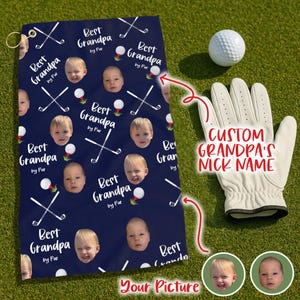 May include: A navy blue golf towel with a repeating pattern of baby faces, golf clubs, and golf balls. The towel has the text "Best Grandpa by Par" in white. A white golf glove and golf ball are also visible.