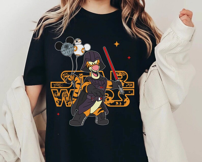 May include: Black t-shirt with a graphic of Tigger dressed as Darth Vader holding a lightsaber. Tigger is holding a Death Star, a TIE fighter, and a Stormtrooper helmet. The text "Star Wars" is printed in orange on the shirt.